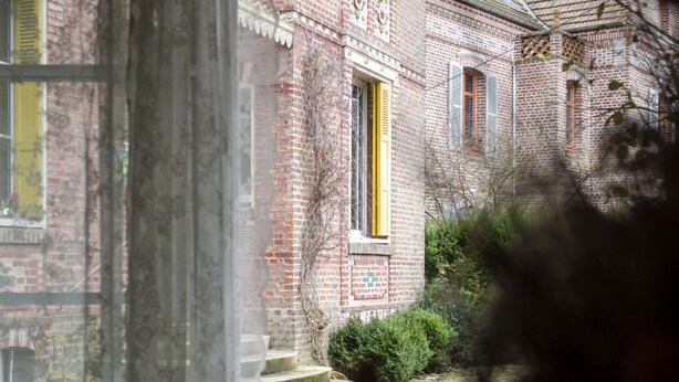 Uitzicht door een raam met gordijn, waarop een bakstenen gebouw met een gele deur en een stenen pad zichtbaar is.