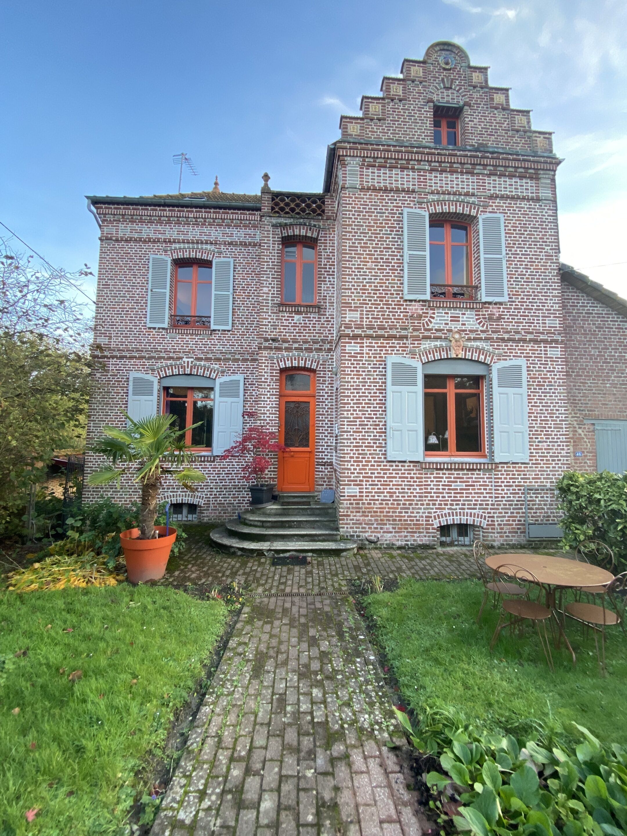 A two-story brick building with a red door and white shutters, a brick pathway leading to the entrance, and a small garden area with a potted plant.