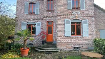 A two-story brick building with a red door and white shutters, a brick pathway leading to the entrance, and a small garden area with a potted plant.