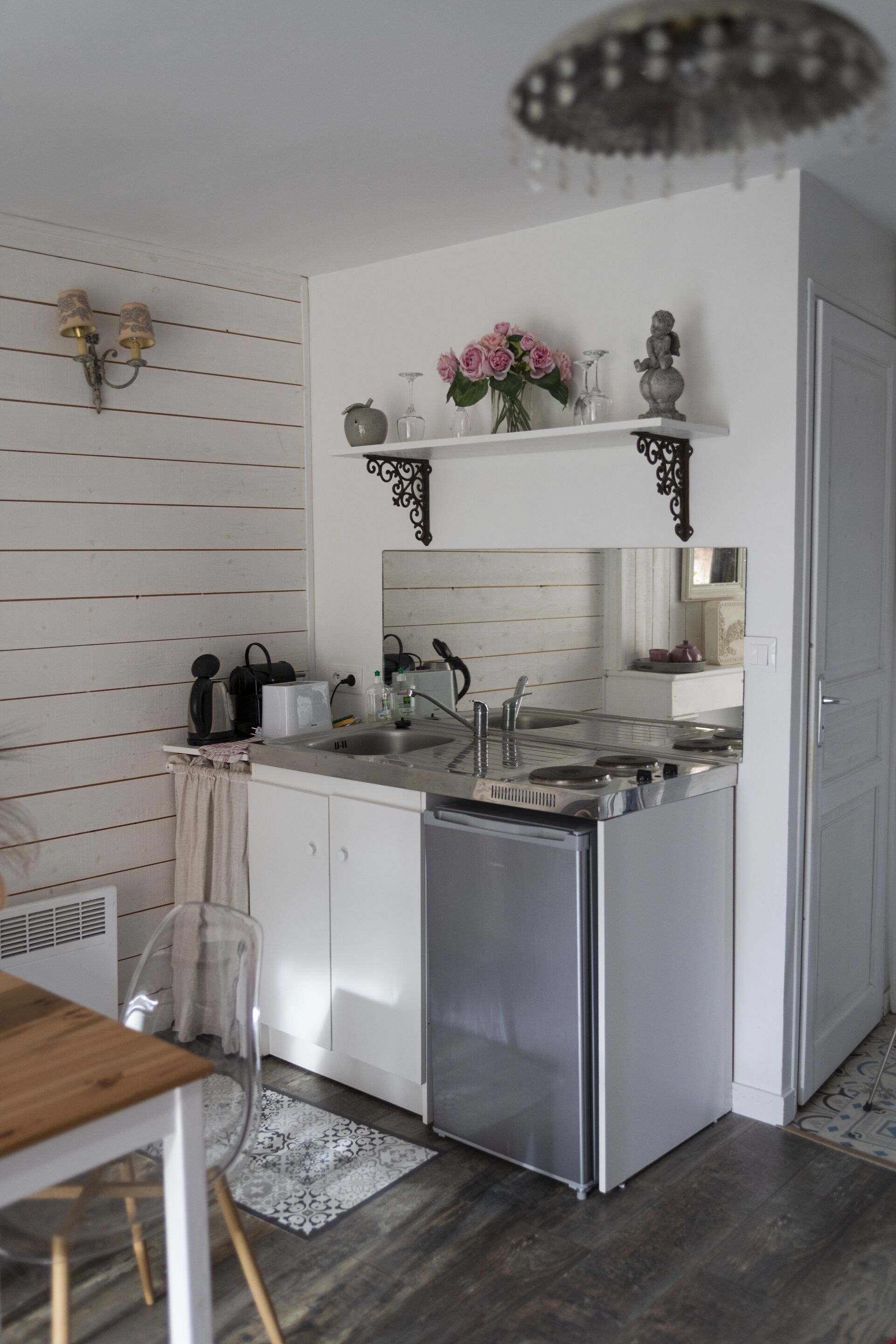 Een compacte keuken met een ingebouwde vaatwasser, een spoelbak en een aanrecht. Boven de spoelbak hangt een decoratieve plank met bloemen en een klein beeldje.