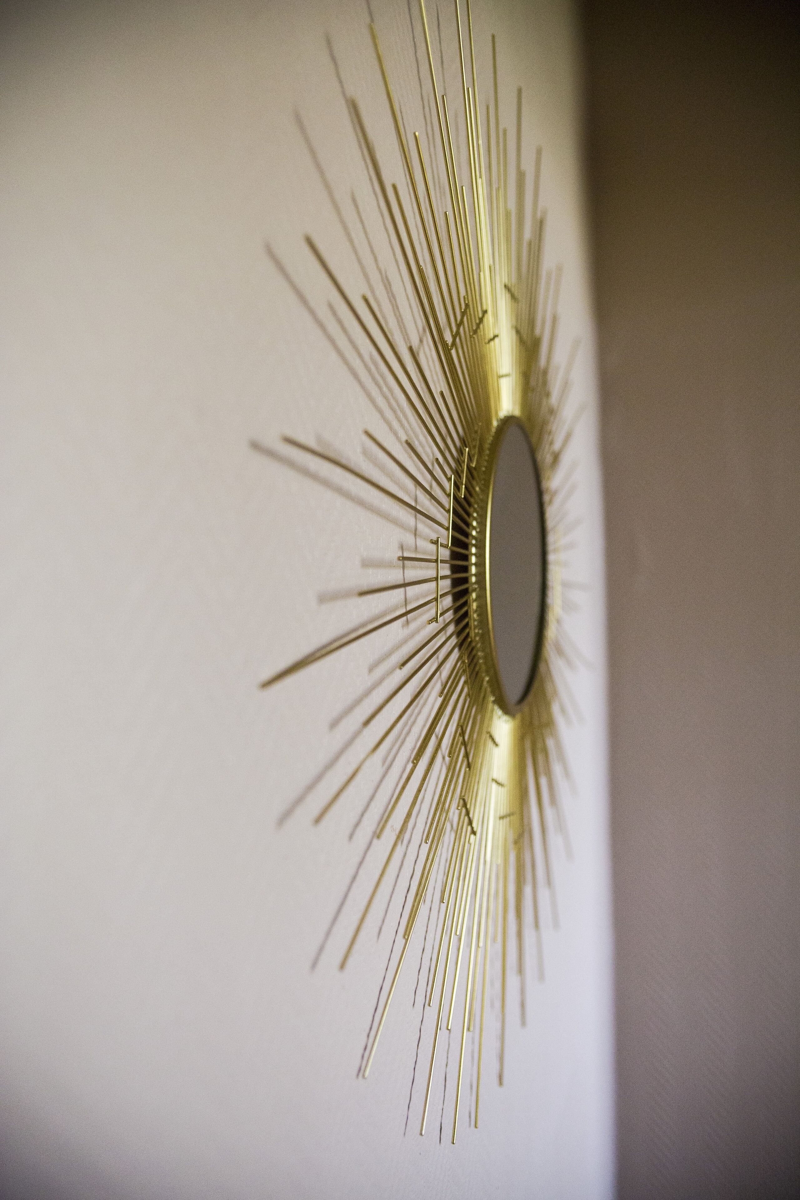 A wall-mounted, sunburst-style mirror with a central oval mirror surrounded by radiating metallic elements.