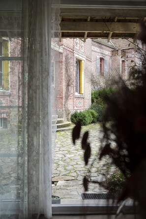 Une vue à travers une fenêtre, à travers un rideau, qui révèle un bâtiment en briques avec une porte jaune et un chemin de pierre menant à une autre porte.