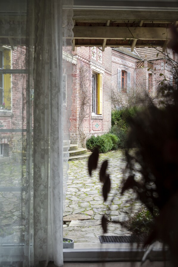 Blick durch ein Fenster mit Vorhang, der ein Backsteingebäude mit gelber Tür und einen gepflasterten Weg zu einer Tür zeigt.