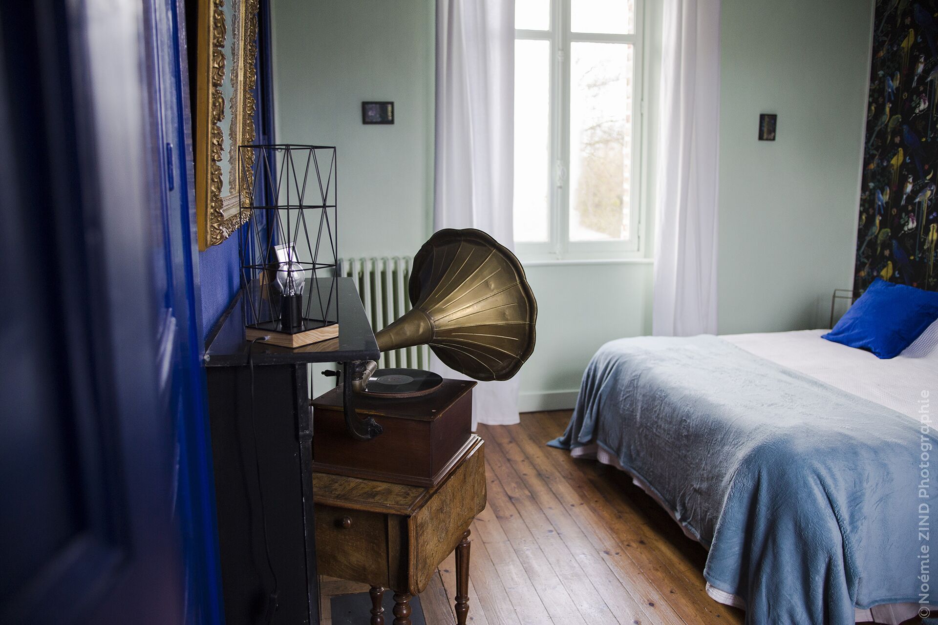 Une chambre à coucher avec un lit, un gramophone ancien, un radiateur et une fenêtre avec des rideaux.