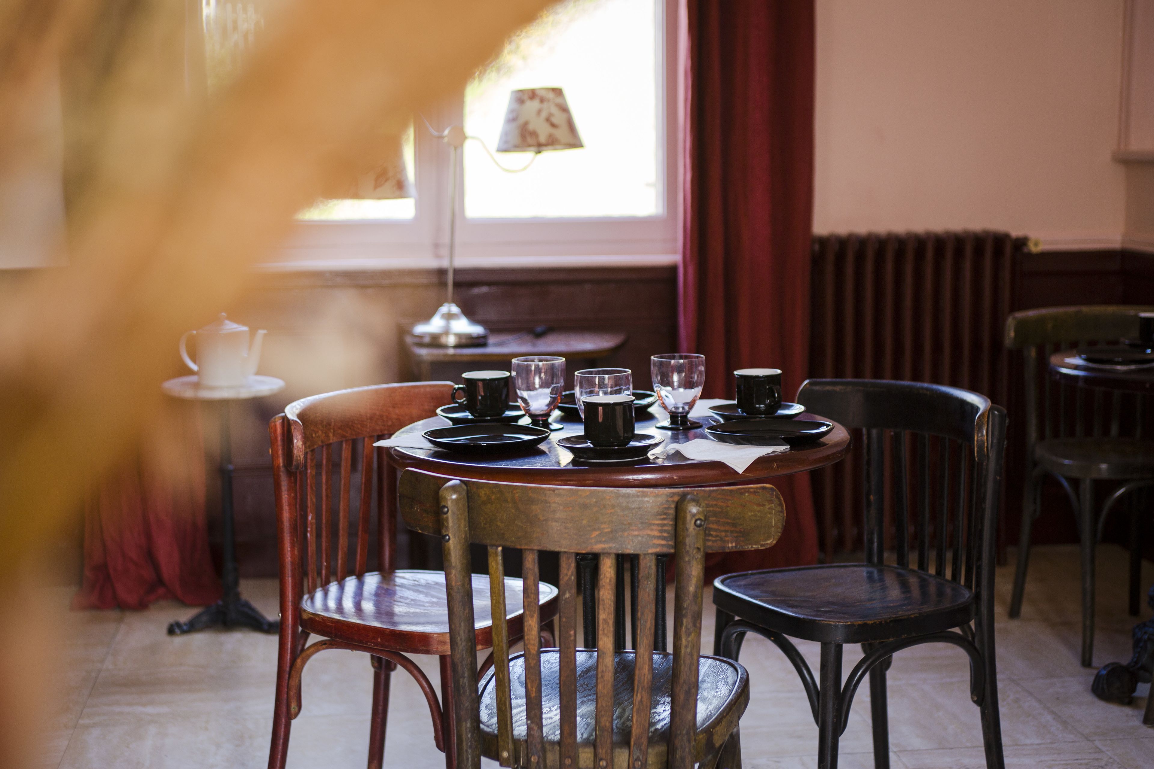 Ein Essbereich mit einem runden Tisch, gedeckt für vier Personen, umgeben von Holzsesseln. Im Hintergrund ist eine Lampe und ein kleiner Tisch mit einem Teekessel zu sehen.
