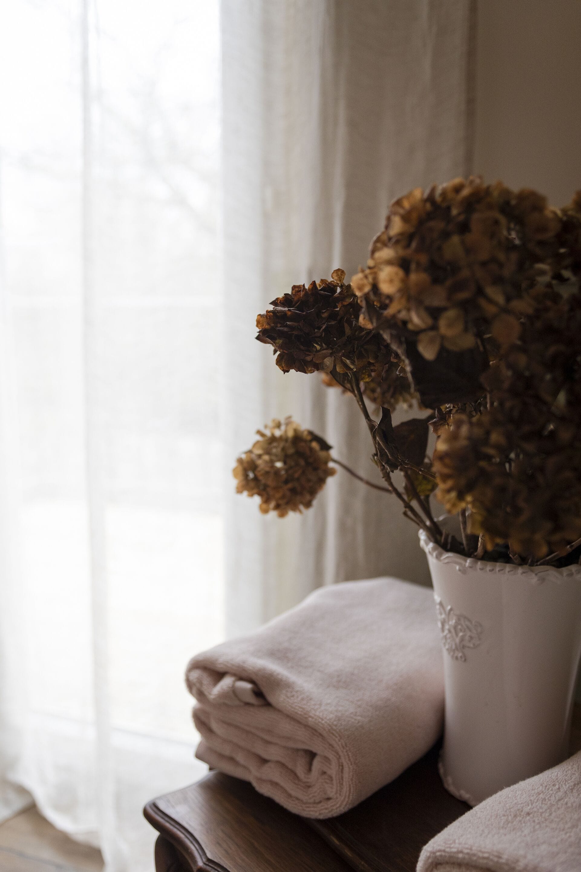 A white vase with dried flowers on a wooden table next to a window with sheer curtains.