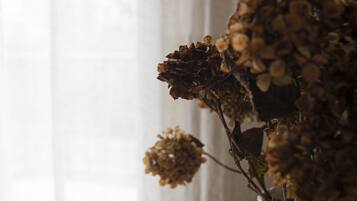 A white vase with dried flowers on a wooden table next to a window with sheer curtains.