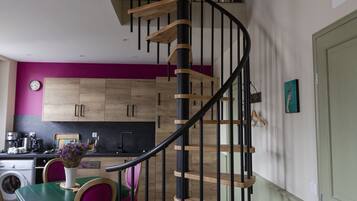 Una cocina moderna con armarios de madera, un comedor con mesa verde y sillas moradas, y una escalera en espiral con barandillas negras.