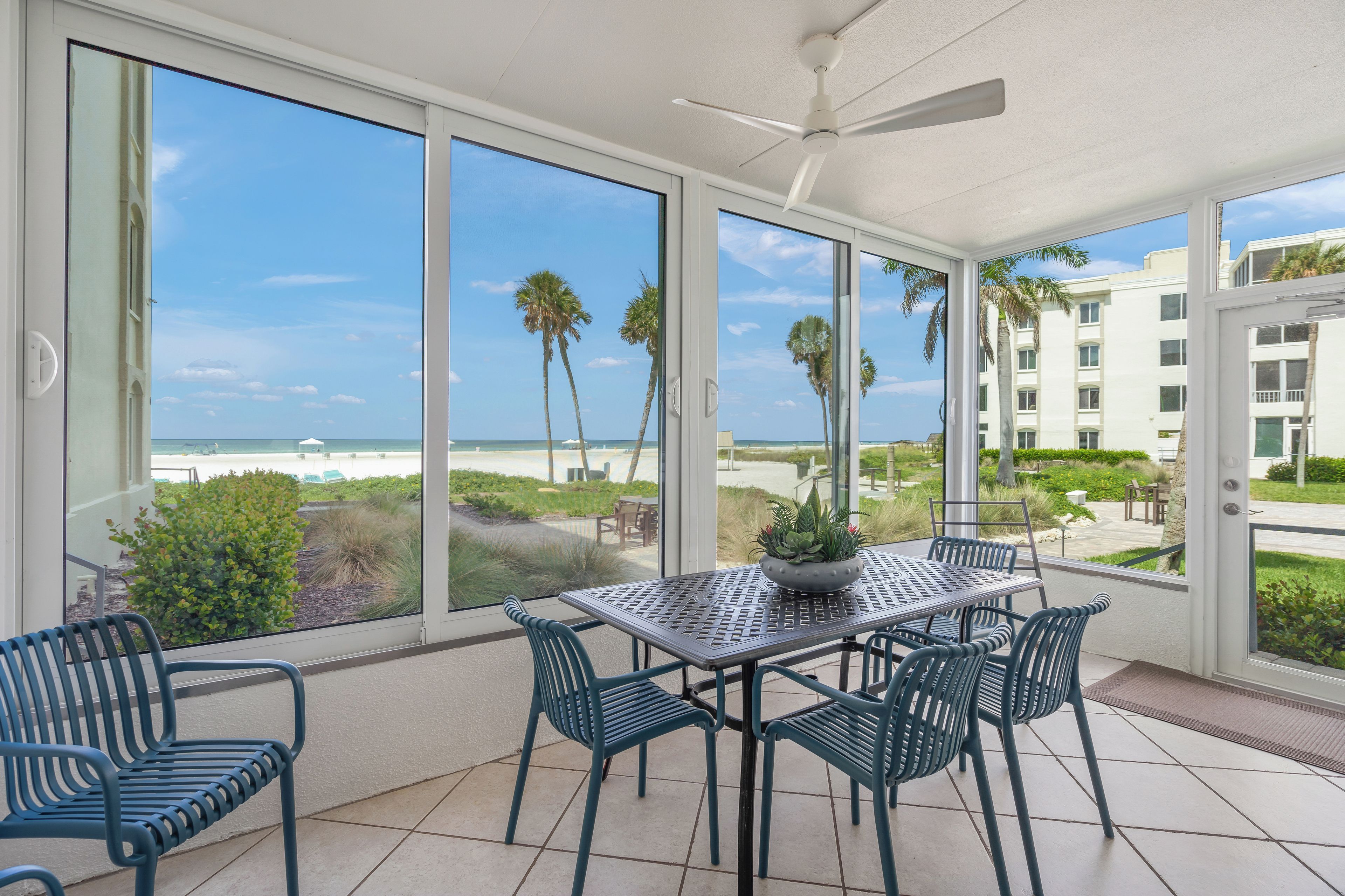 A sunlit room with a view of the beach, a table with chairs, and a potted plant.