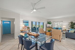 A spacious living area with a glass dining table, blue chairs, a ceiling fan, and a view of the outdoors through large windows. - Island House Beach Resort 2S (Siesta Key)