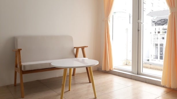 Habitación con un sofá blanco y una mesita pequeña de patas de madera, una ventana grande con cortinas y vistas a edificios.