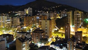 Paisagem urbana noturna com edifícios altos, uma colina notável ao fundo e postes de luz iluminando a área.