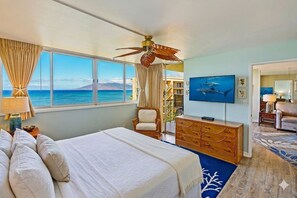A hotel room with a bed, a ceiling fan, a TV, a dresser, and a view of the ocean.