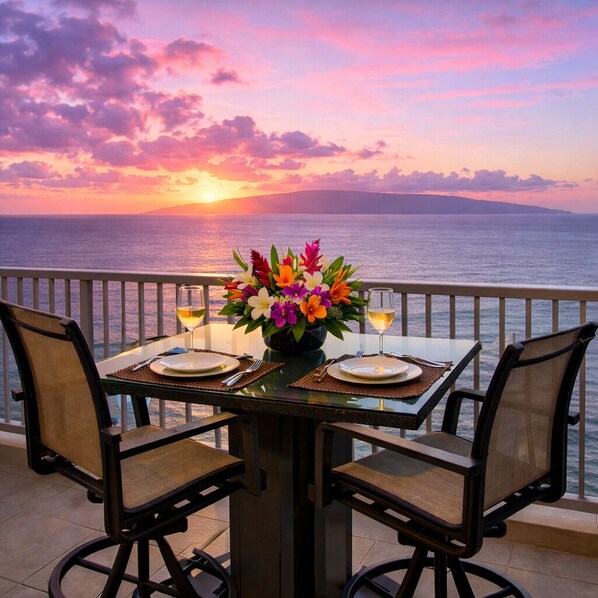 A dining table with chairs, a vase of flowers, and two glasses of wine on a balcony overlooking the sea at sunset.