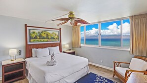 A hotel room with a bed, a nightstand, a chair, a ceiling fan, a window with a view of the ocean, and a painting on the wall.