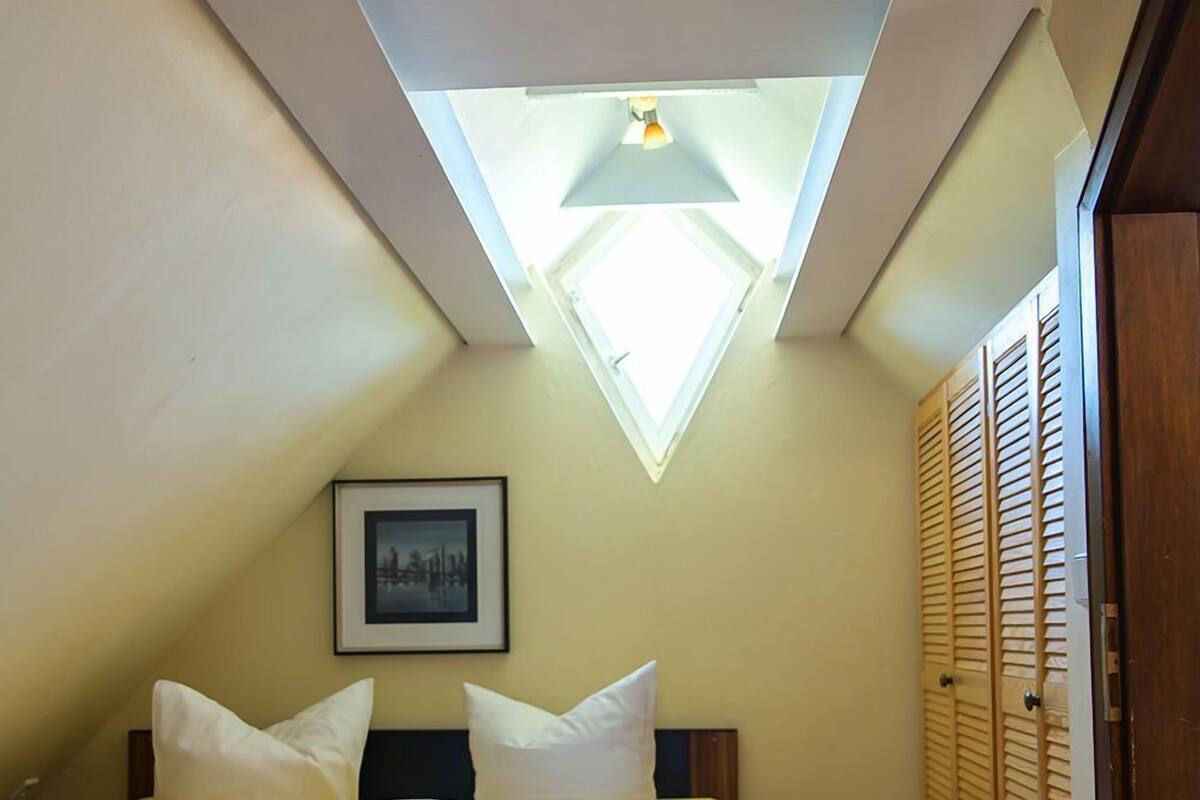 Une chambre avec une lucarne, un lit avec des oreillers, un tableau encadré au mur et un plancher en bois.
