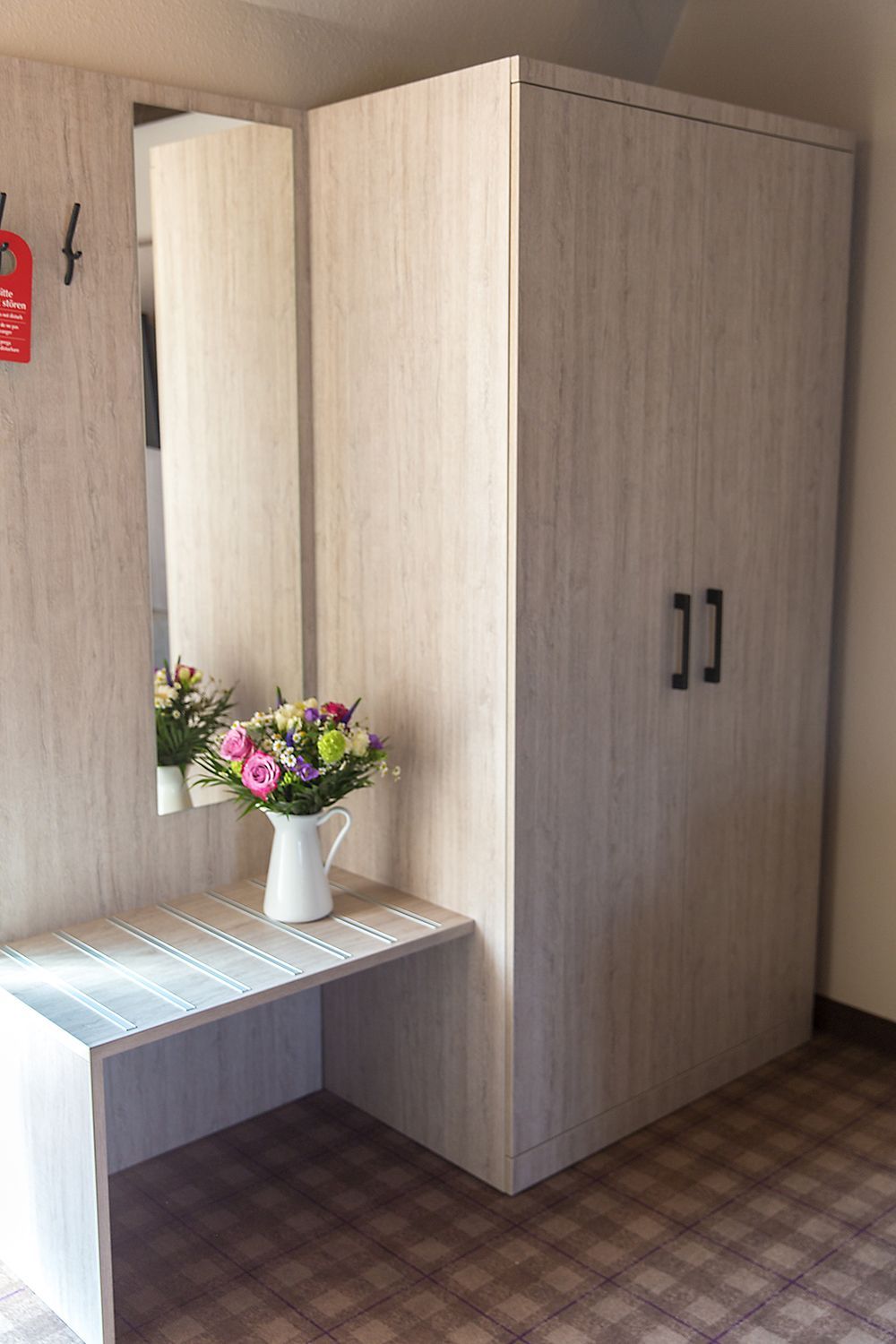 A room with a wardrobe, a mirror, and a small table with a vase of flowers.