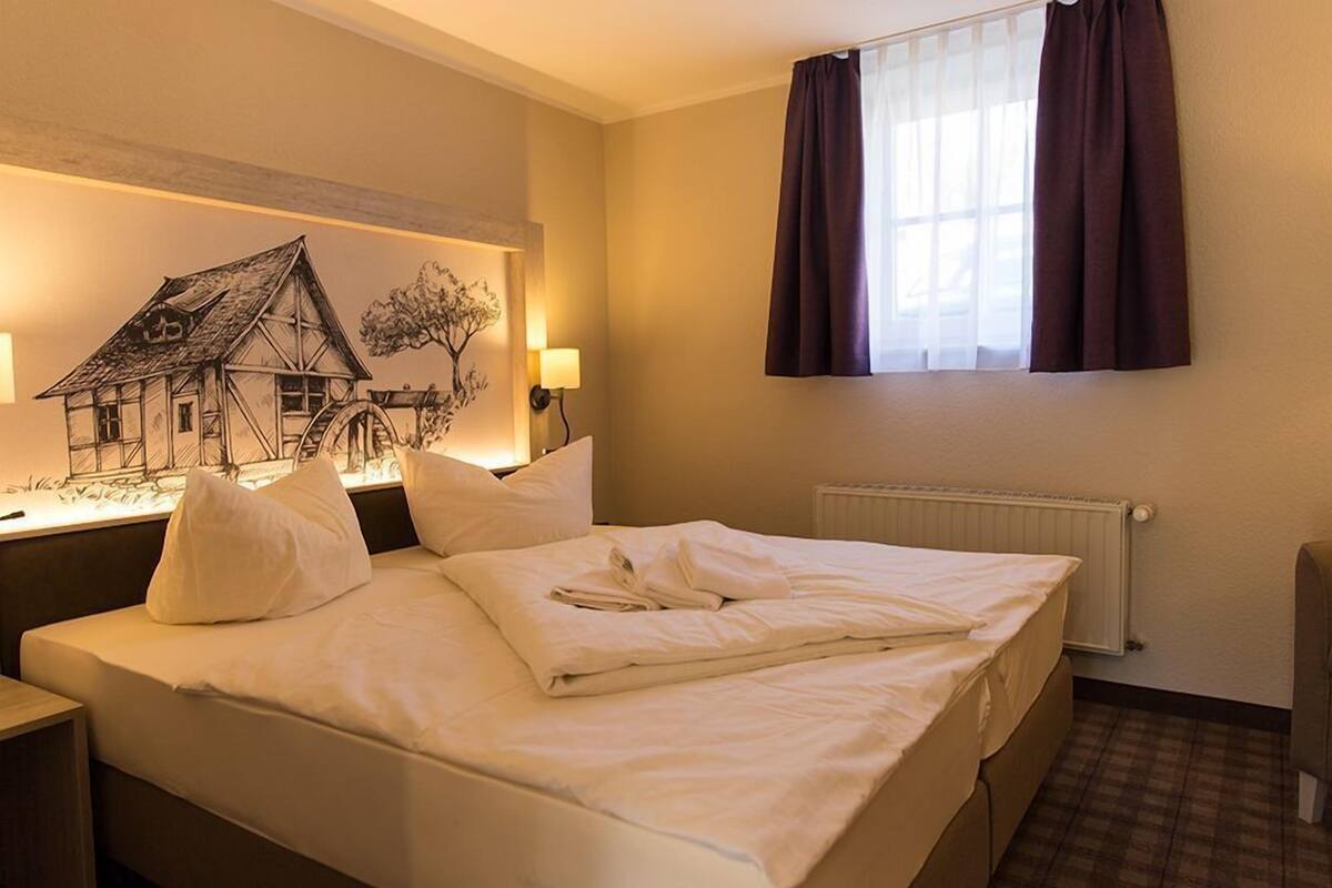 Une chambre d’hôtel avec un lit, une table de chevet, une lampe, une fenêtre avec des rideaux et une œuvre d’art représentant une maison, accrochée au mur.