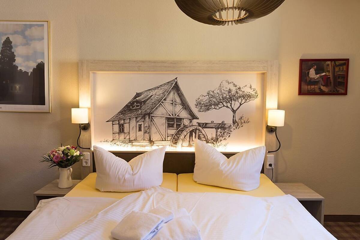 Un lit bien fait, avec du linge de lit blanc et des oreillers, une œuvre d’art représentant une maison et un arbre accrochée au mur, et deux lampes de chevet.