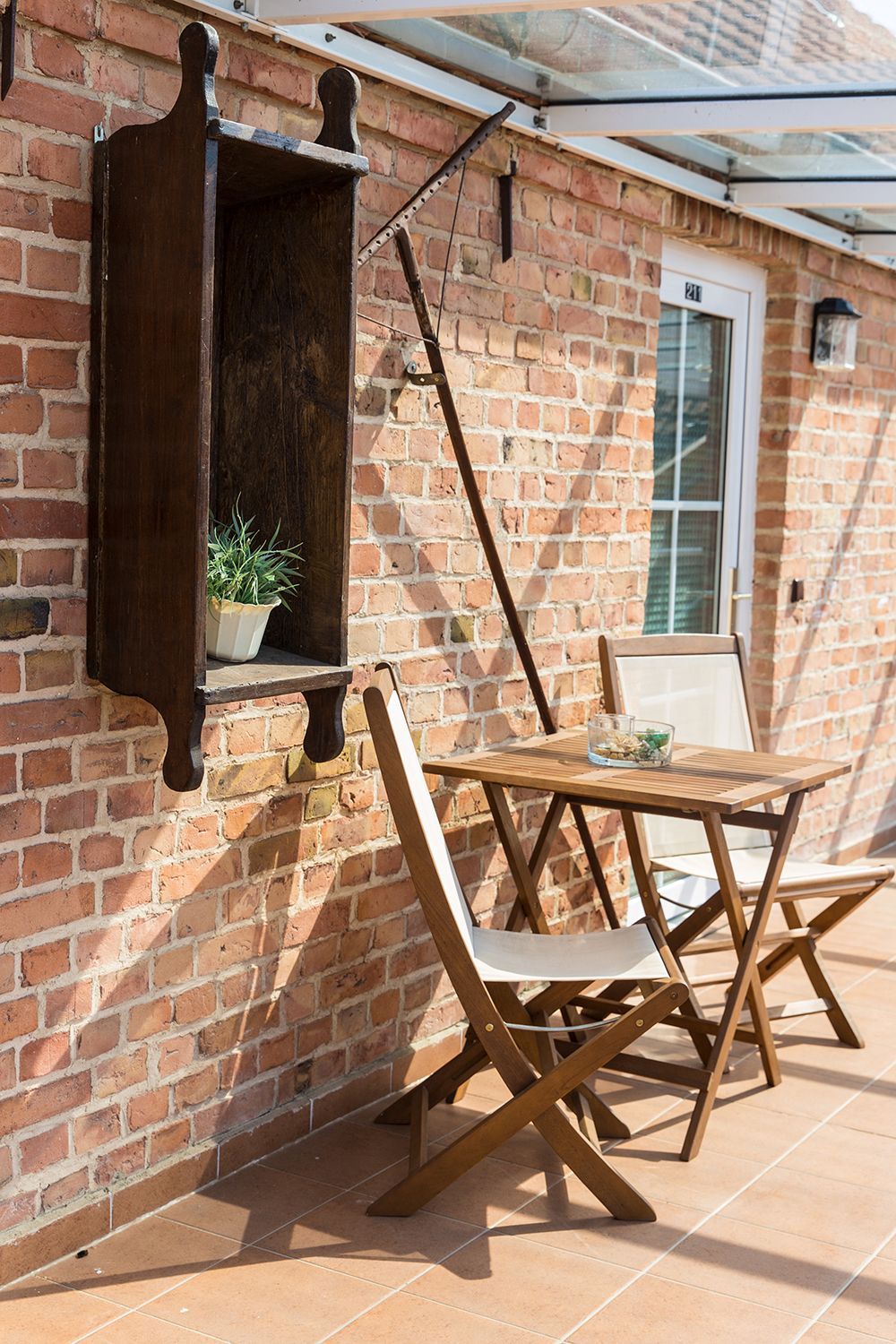 A brick wall with a wooden table and chairs, a hanging shelf with a plant, and a window with a white frame.
