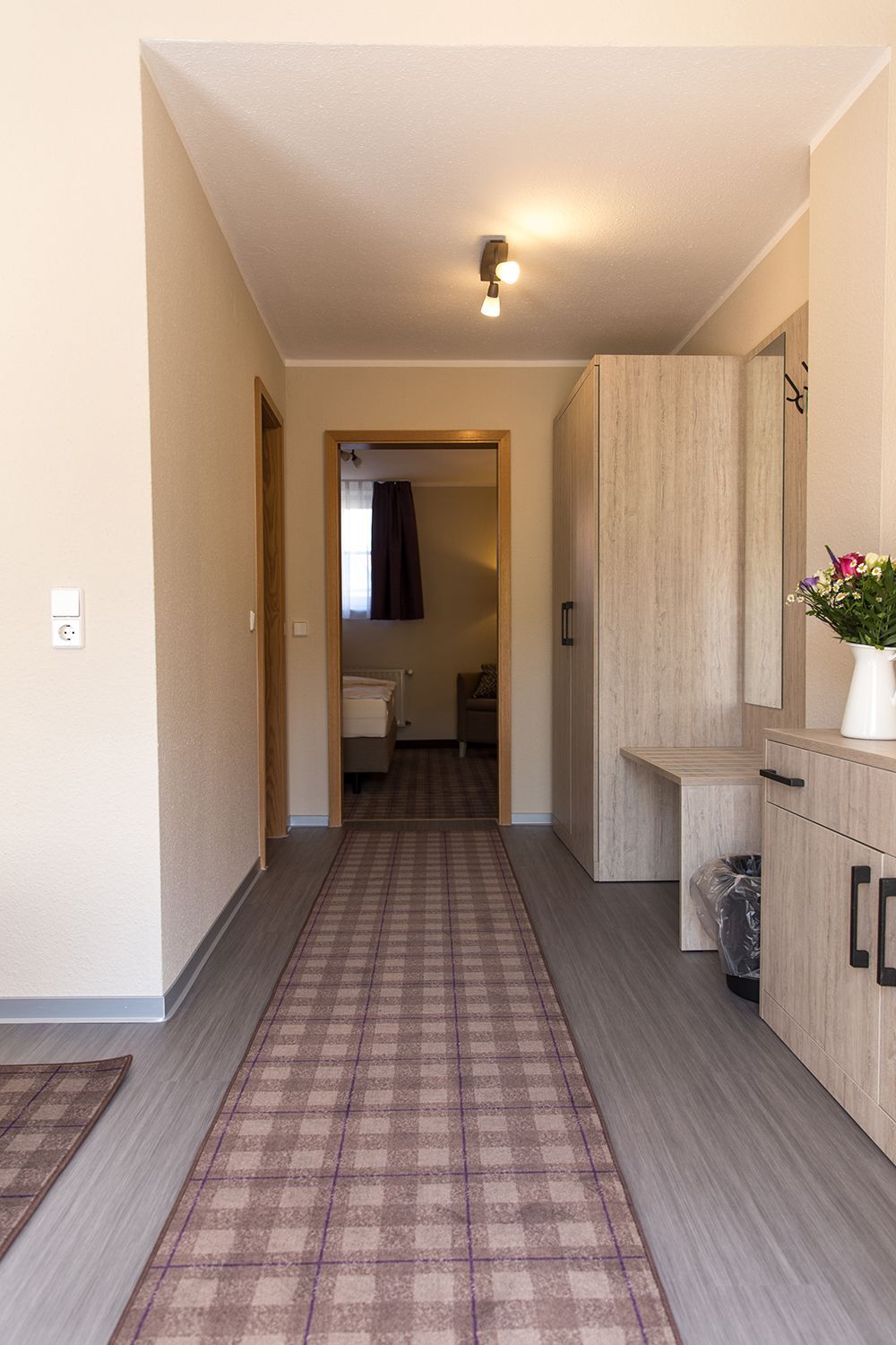 A hallway with a checkered carpet, wooden cabinets, a light fixture, and a door leading to a bathroom.