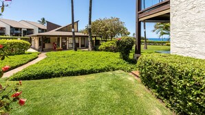 A well-maintained garden with manicured lawns and hedges, a building with a modern architectural style, and a clear view of the ocean in the background. - Hale Kamaole #107 Ground floor condo, easy access to best beach on Maui! (Kihei)