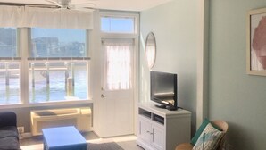 A living room with a ceiling fan, a television on a white cabinet, a window with curtains, a wooden table, and a chair.