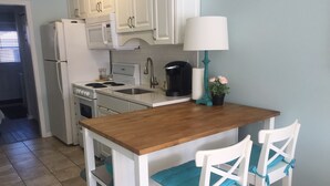 A compact kitchen with white cabinets, a wooden countertop, a sink, a stove, a microwave, and a coffee maker. There are two white chairs with blue cushions and a teal lamp.