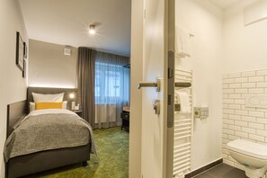 Een hotelkamer met een bed, nachtkastje, lamp, raam met gordijnen en een badkamer met toilet en handdoekhouder.
