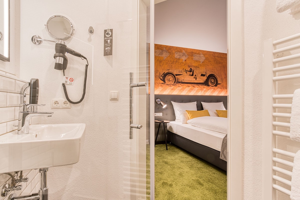 Une chambre d’hôtel dotée d’une salle de bain comprenant un lavabo, un miroir et un sèche-cheveux. La chambre est équipée d’un lit, d’une moquette verte et d’un tableau représentant une voiture accroché au mur.