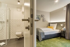 Een hotelkamer met een douche, toilet, bed en een klein tafeltje met een lamp.