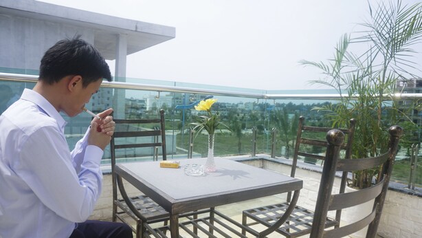 A man sitting at a table with a vase of yellow flowers, a glass of water, and a nameplate on a balcony with a city view.