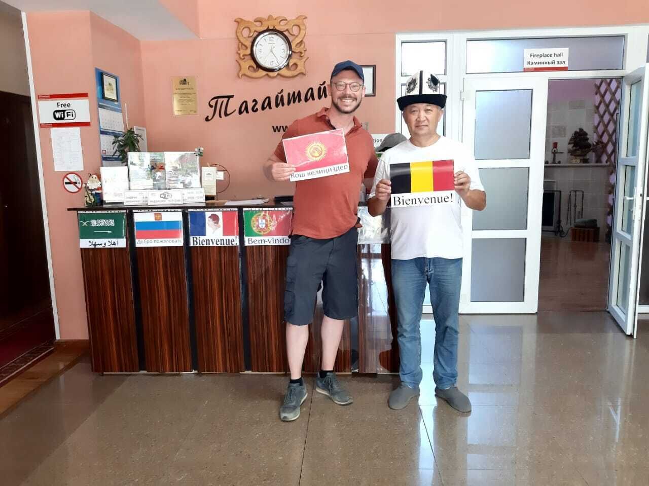 Two men holding certificates in a reception area with a clock, a sign for the fireplace hall, and multiple language welcome signs.