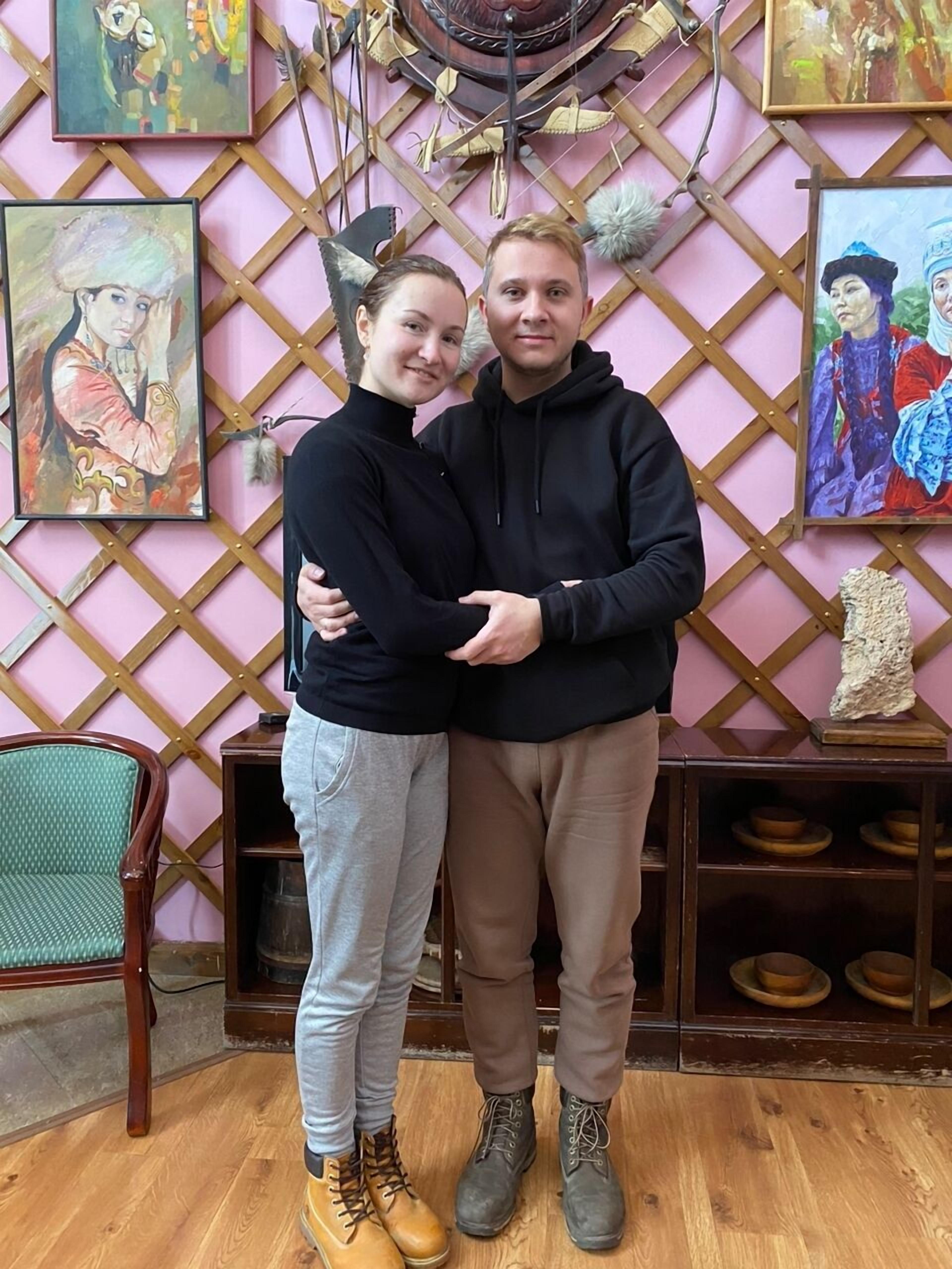 A couple embracing in a room with wooden floors, a green chair, and artwork on the walls.