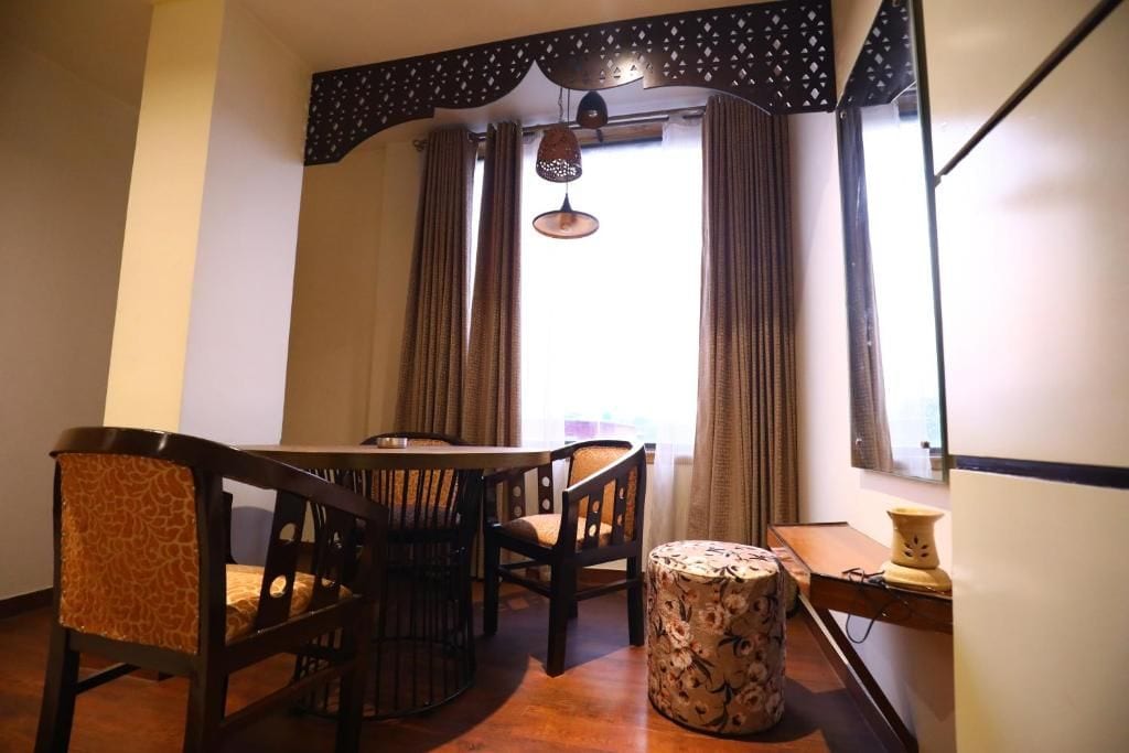 A dining area with a round table, chairs, a patterned ottoman, and a window with curtains.