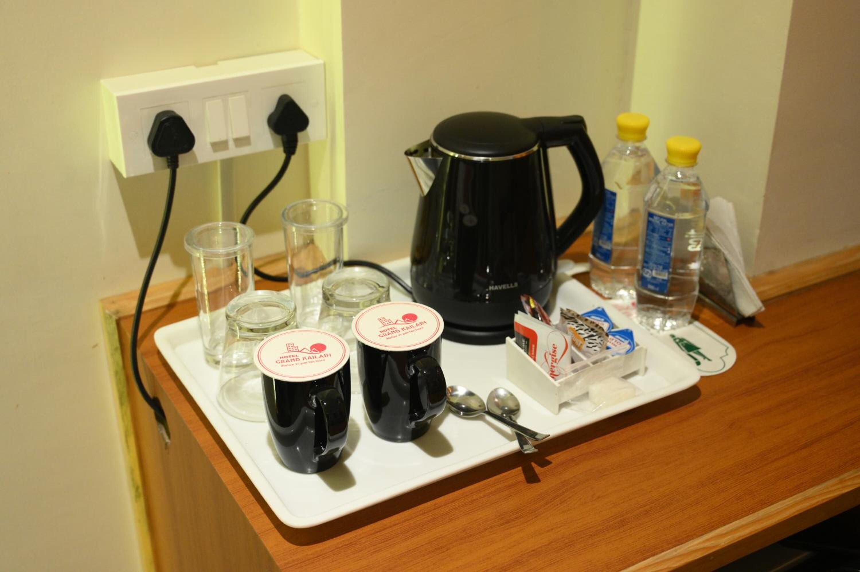 A tray with a coffee maker, two mugs, glasses, a bottle of water, a tissue box, and a spoon on a wooden counter.