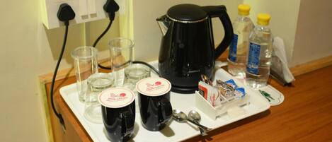 A tray with a coffee maker, two mugs, glasses, a bottle of water, a tissue box, and a spoon on a wooden counter.