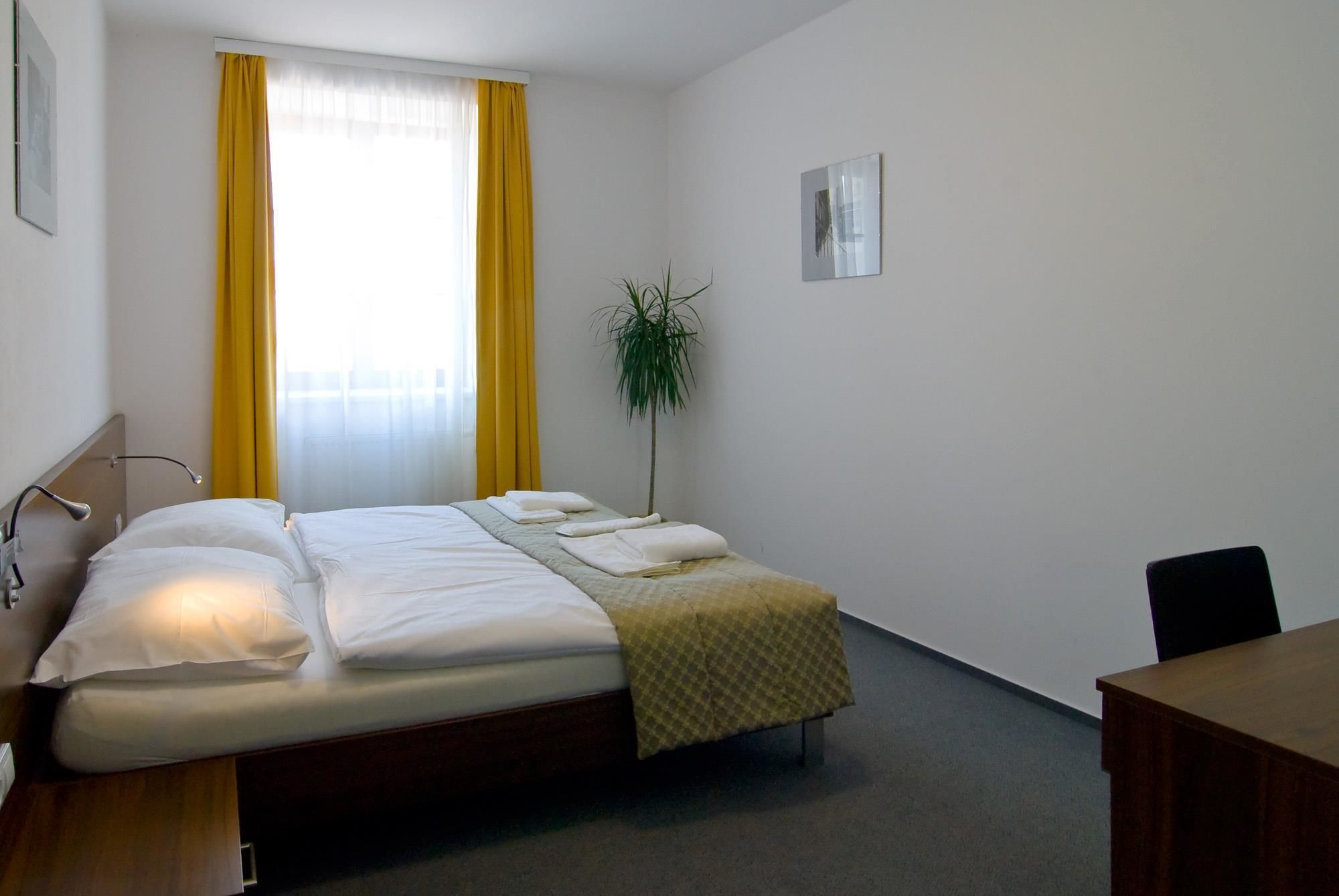 Une chambre d’hôtel avec un lit double, un bureau, une chaise, une plante et une fenêtre avec des rideaux jaunes.