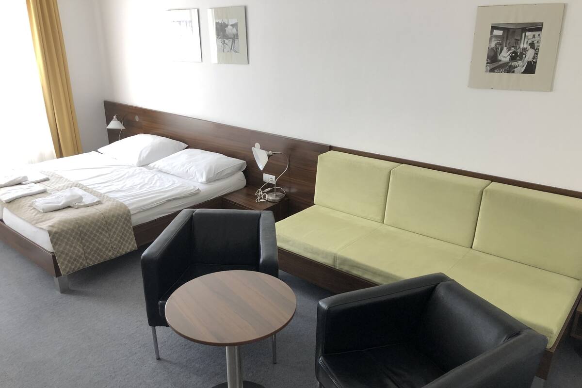 Una habitación de hotel con una cama, un sofá, dos sillones, una mesita redonda y una mesita de noche con lámpara.
