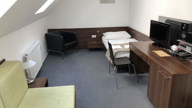 Habitación de hotel compacta con cama, escritorio con computadora, silla, lámpara y radiador.