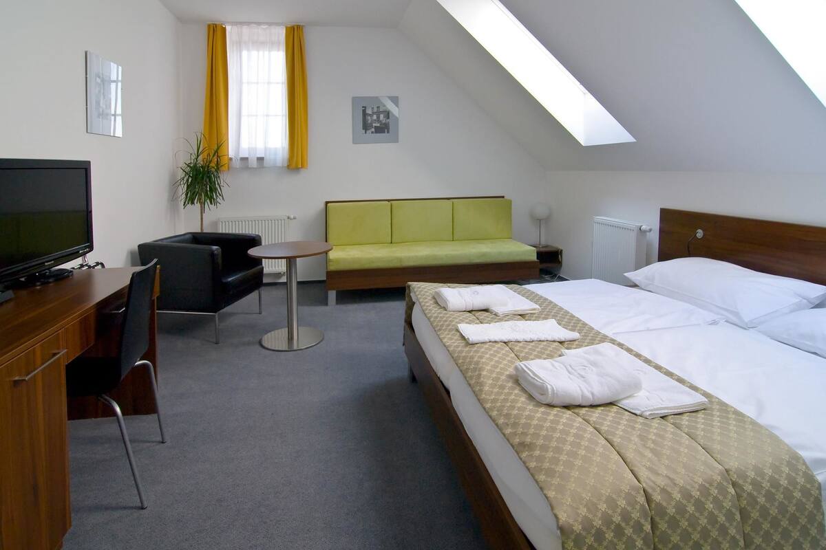 Habitación de hotel con cama, escritorio, silla, mesita, sofá, televisión y ventana con cortinas.