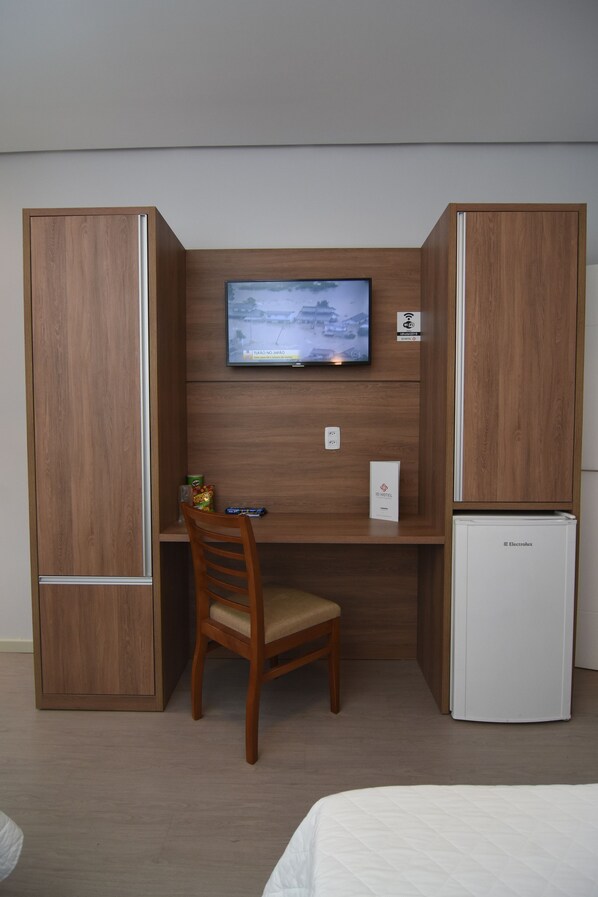 A compact room with a wooden desk, a television mounted on the wall, a chair, and a small refrigerator.