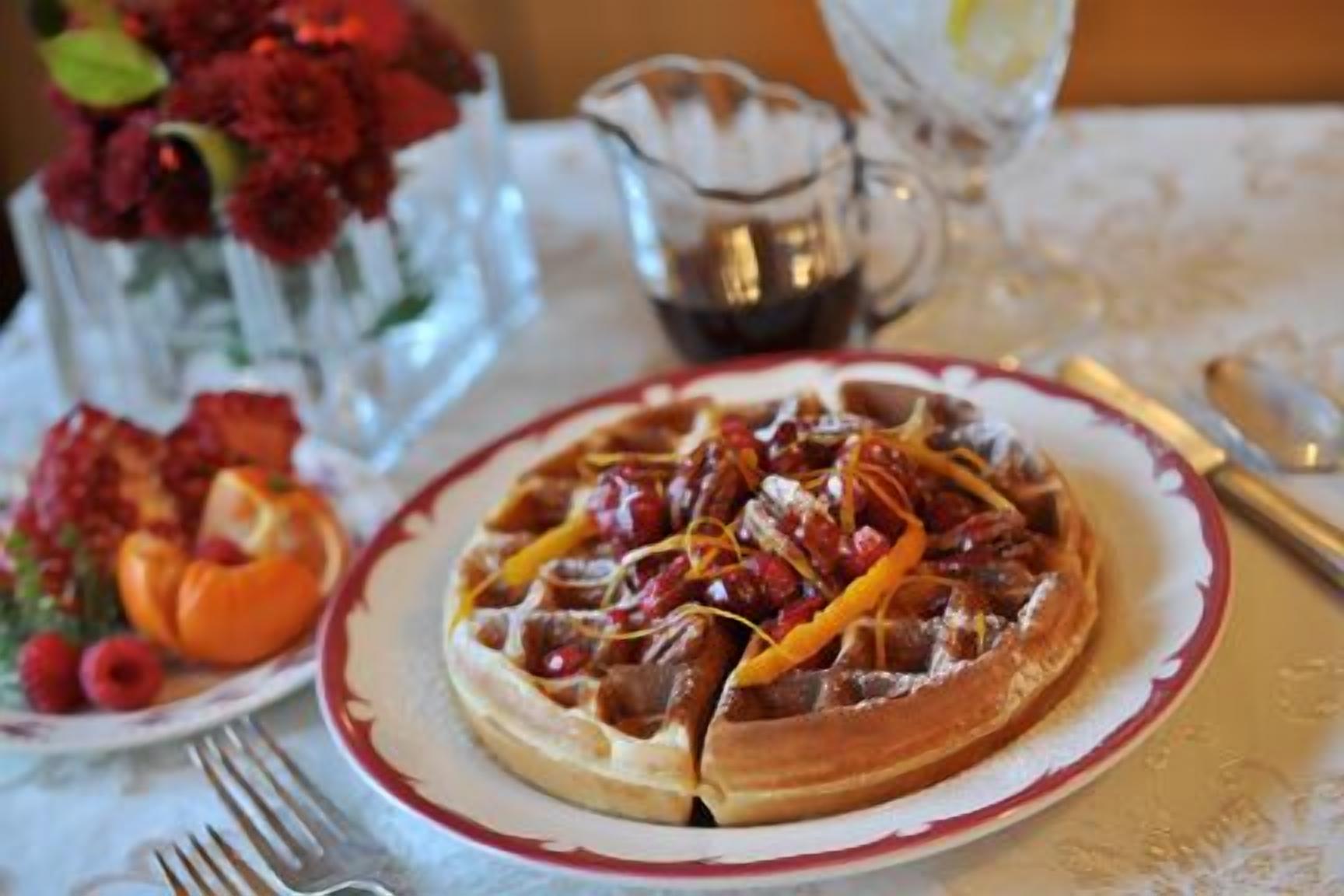 A plate of food with a waffle, fruit, and a pitcher of syrup on a table.
