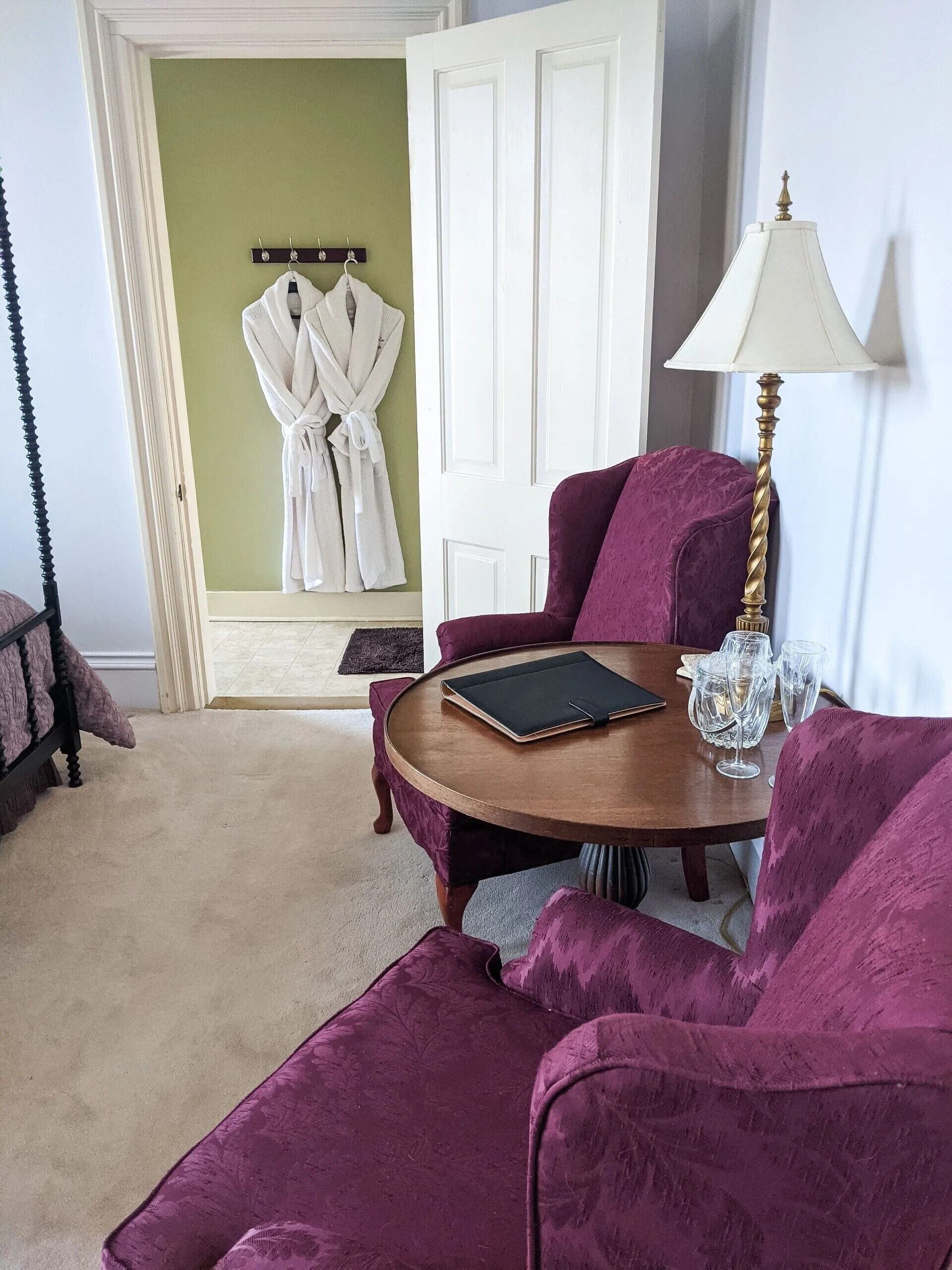 A room with a round wooden table, two purple armchairs, a white door, and a robe hanging on a hook.