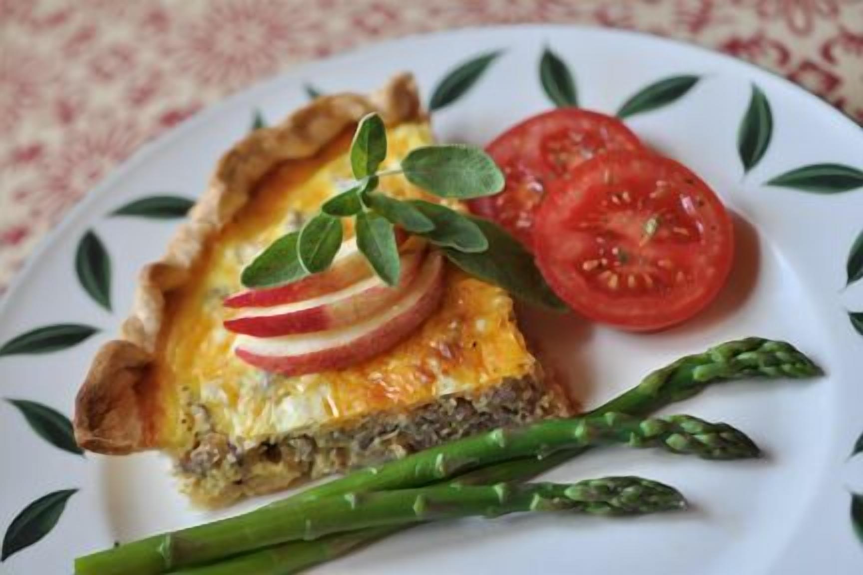 A slice of quiche with a crust, garnished with apple slices and a sprig of herb, served with a side of asparagus and a slice of tomato.