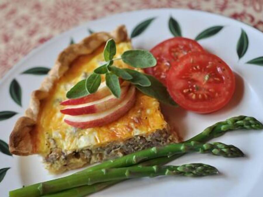 A slice of quiche with a crust, garnished with apple slices and a sprig of herb, served with a side of asparagus and a slice of tomato.