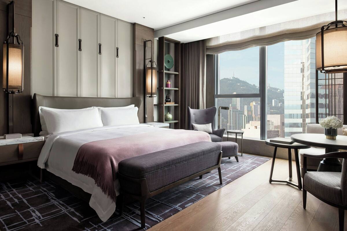 A modern hotel room with a large bed, a seating area, a dining table, and a view of the city.