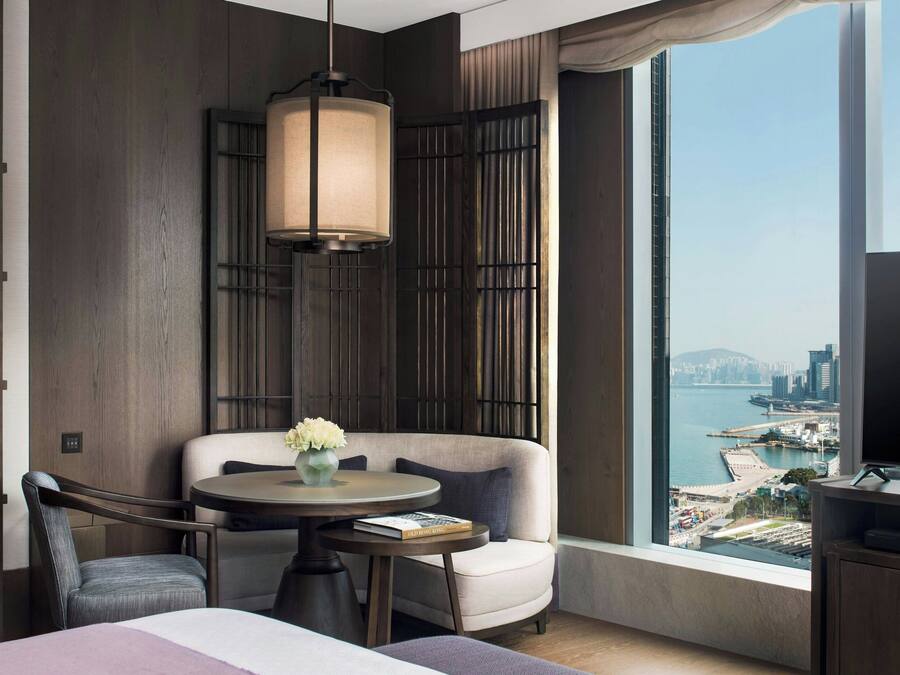 A modern hotel room with a large window offering a city view, a round table with a vase of flowers, a sofa, and a bed.
