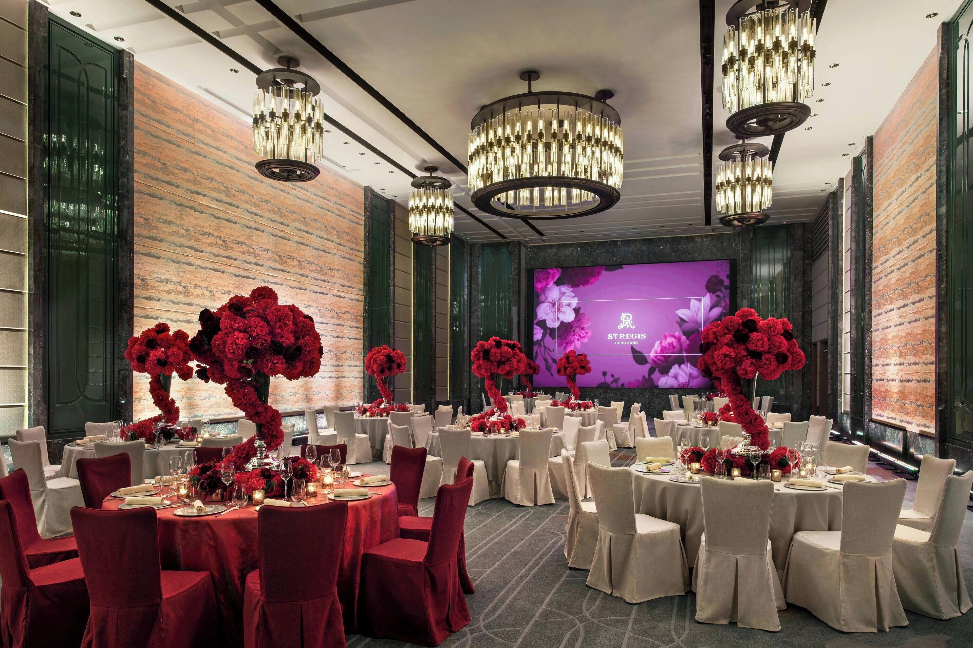 A banquet hall with round tables, white chairs, and red floral centerpieces.