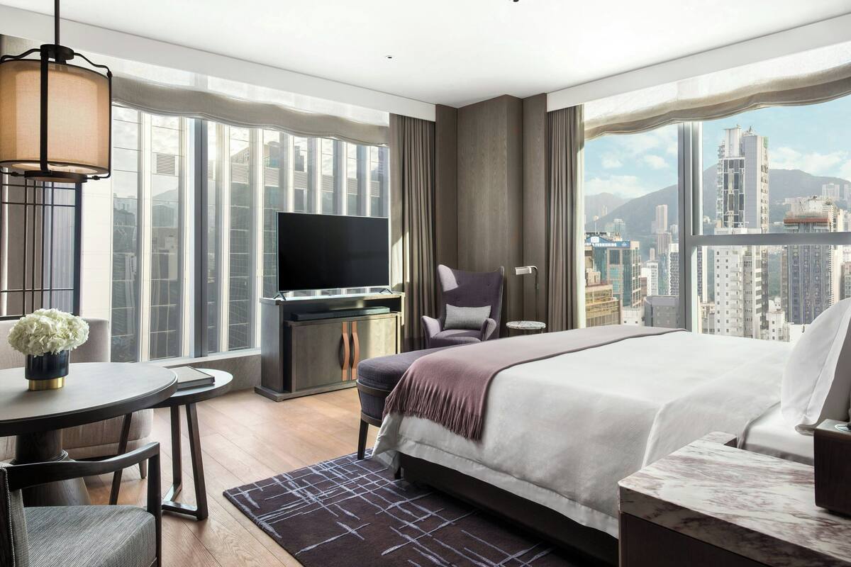 A modern hotel room with a large bed, a desk with a TV, a chair, and a view of the cityscape.